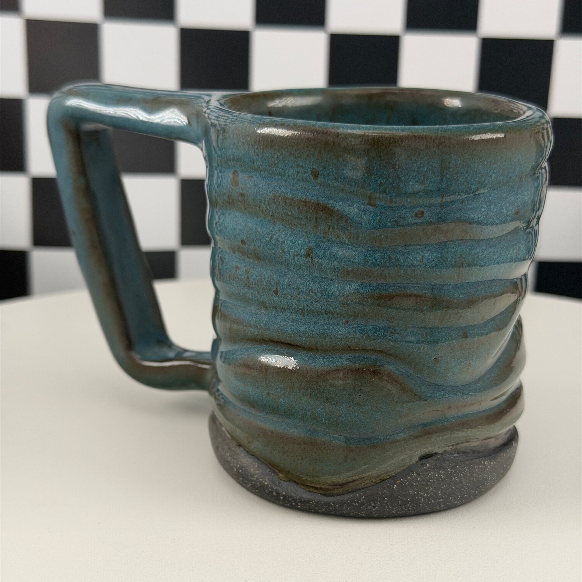A handmade ceramic mug with a wavy, ribbed texture and a glossy teal-blue glaze, set against a black and white checkered background on a white surface.