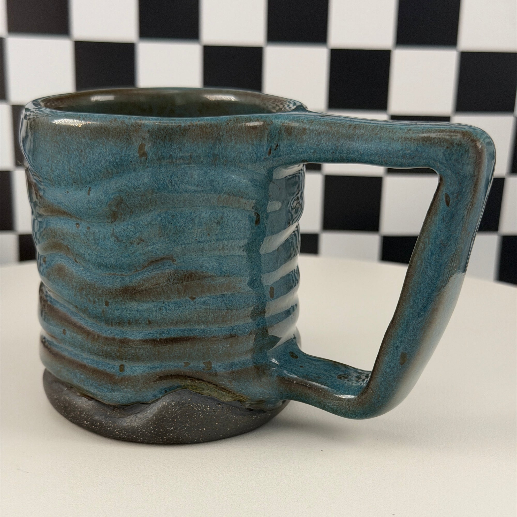 A handmade ceramic mug with a rounded handle and ribbed body, featuring a textured blue-green glaze and a dark brown base, shown against a white background with a black-and-white checkered scale pattern on the left.
