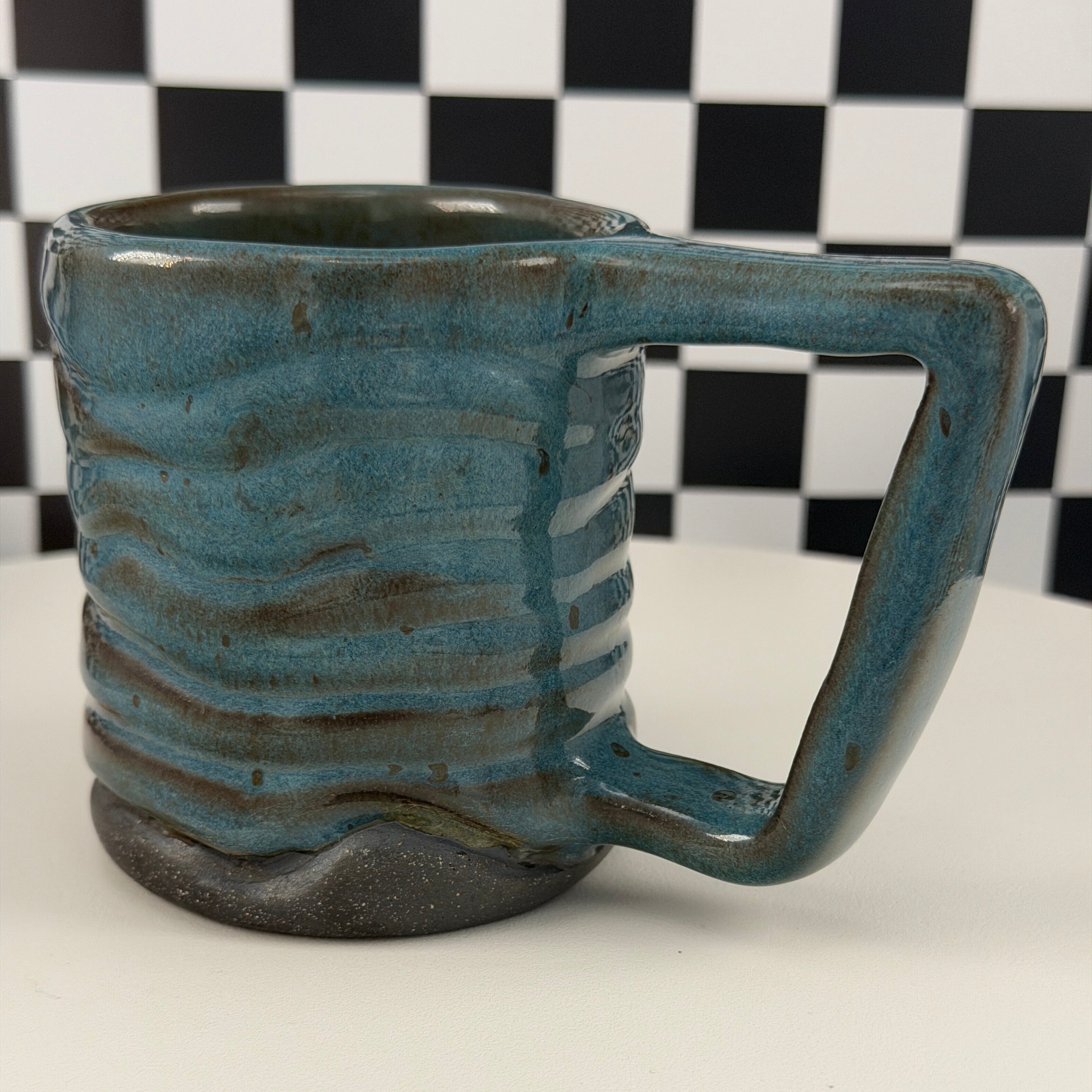 A handmade ceramic mug with a rounded handle and ribbed body, featuring a textured blue-green glaze and a dark brown base, shown against a white background with a black-and-white checkered scale pattern on the left.