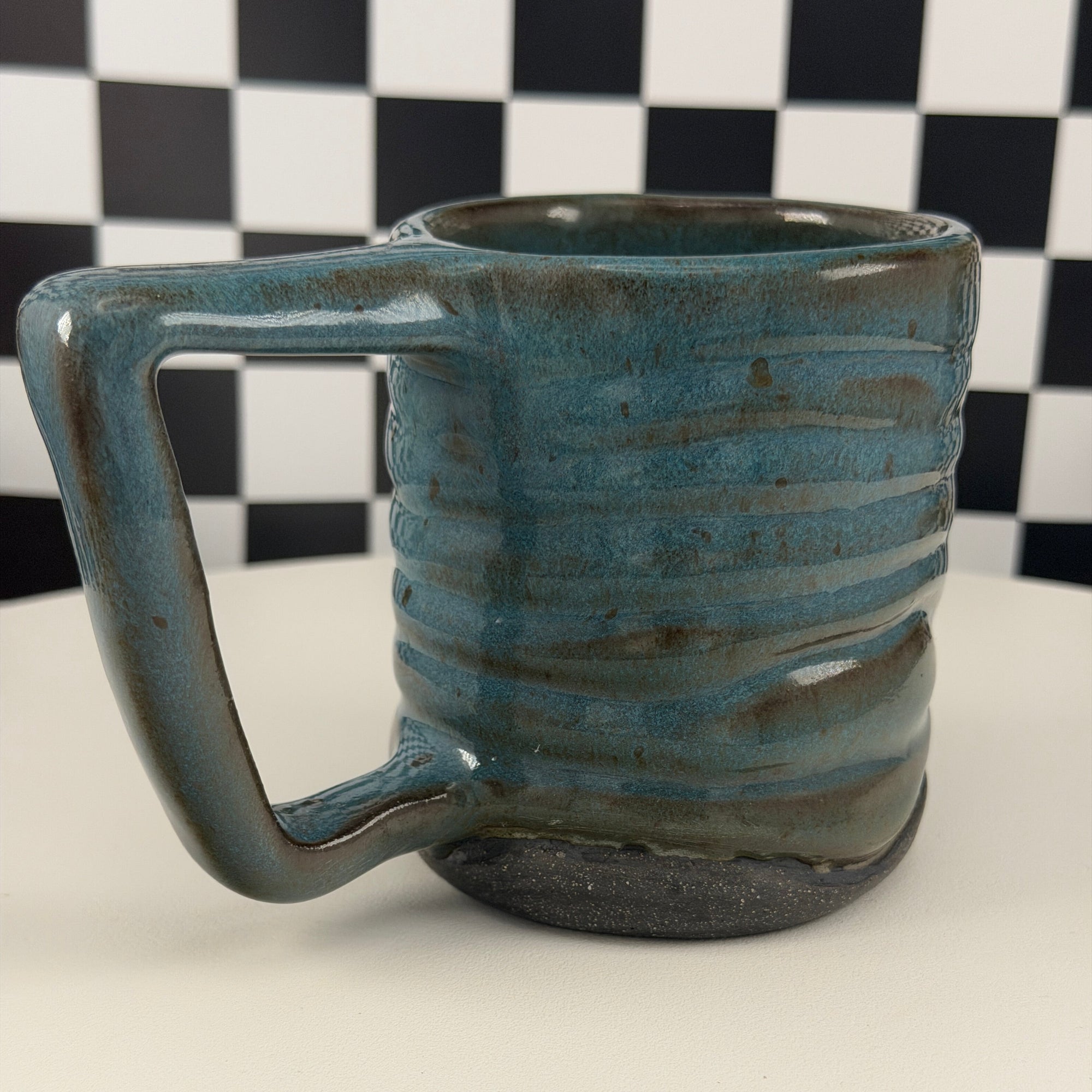 A handmade ceramic mug with a rustic handle, featuring a textured glaze in shades of teal blue and brown, set against a black and white checkered background.
