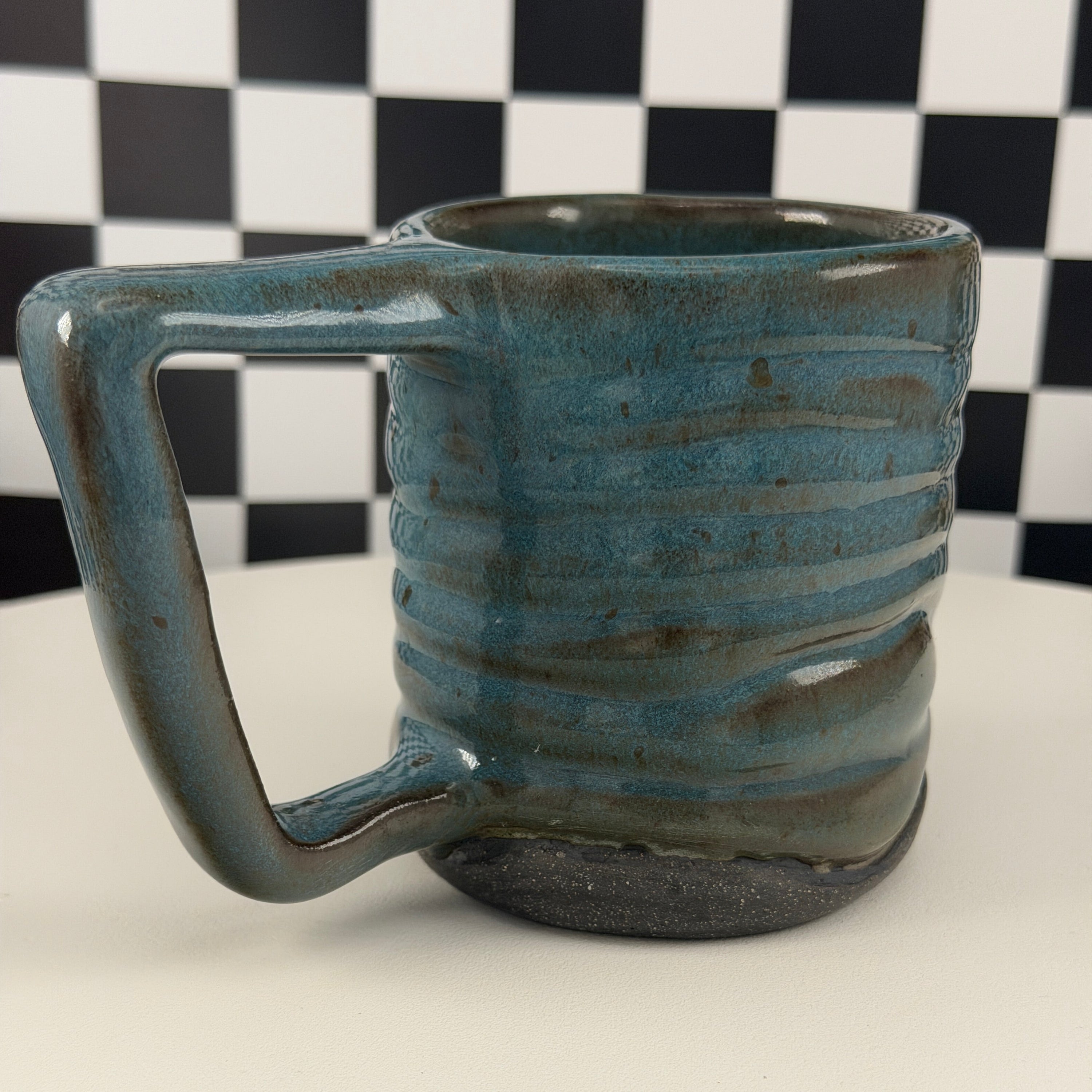 A handmade ceramic mug with a rustic handle, featuring a textured glaze in shades of teal blue and brown, set against a black and white checkered background.